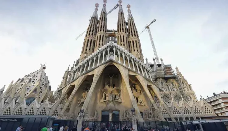A low-angle photograph of the intricate stone towers of Antoni Gaudi's Sagrada Família basilica against a bright blue sky.