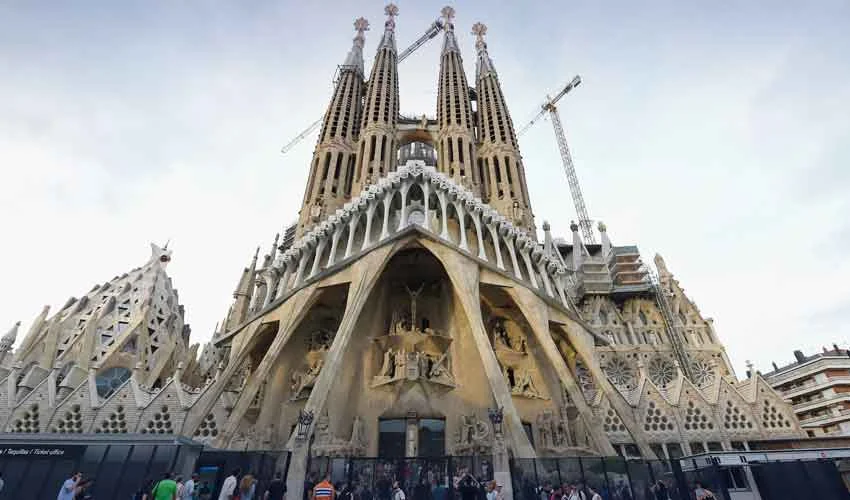 A low-angle photograph of the intricate stone towers of Antoni Gaudi's Sagrada Família basilica against a bright blue sky.
